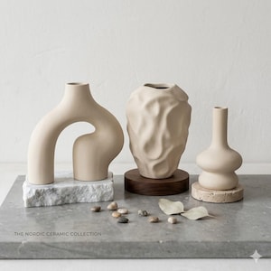 Peut inclure: Une collection de vases en céramique aux tons neutres et aux formes uniques. L'un des vases a un design en arche, un autre une surface texturée et le troisième une forme bulbeuse. Les vases sont présentés sur des socles en marbre, en bois et en pierre, avec des galets et une feuille.