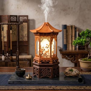 Puede incluir: Un difusor de madera con forma de pagoda que emite una columna de niebla blanca. La estructura presenta un techo claro, un panel central iluminado con motivos florales naranjas y una base hexagonal oscura tallada sobre una bandeja de piedra.