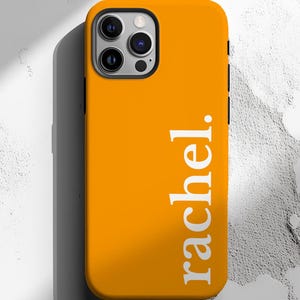 May include: Bright orange phone case with the name "rachel." printed vertically in white. The case has a black border around the camera and sides. The phone case is on a textured white surface.