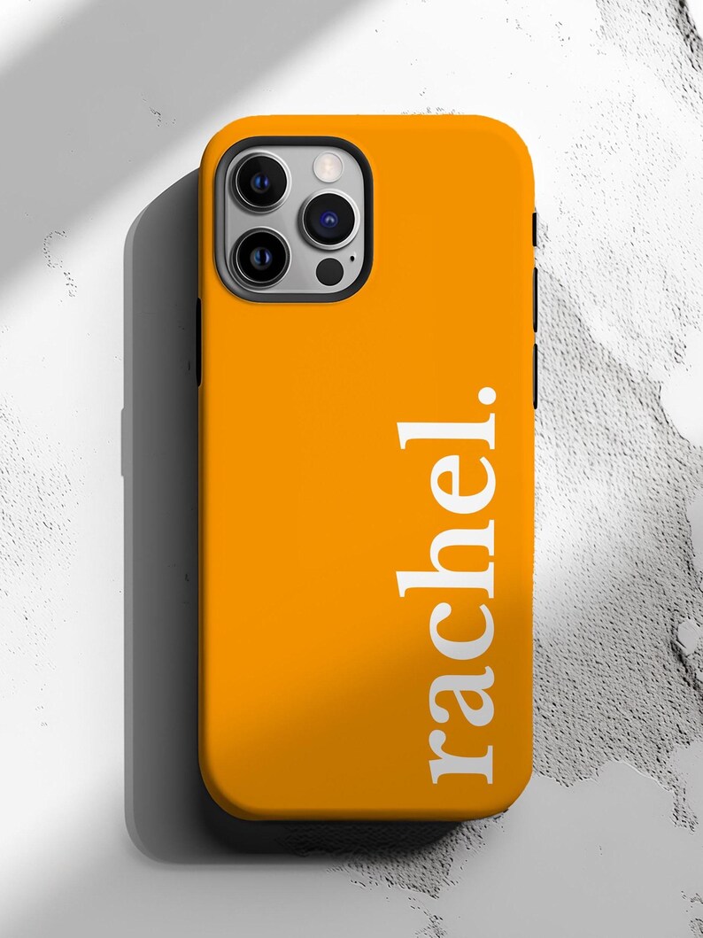 May include: Bright orange phone case with the name "rachel." printed vertically in white. The case has a black border around the camera and sides. The phone case is on a textured white surface.