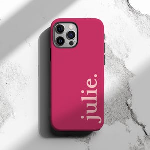 May include: A magenta phone case with the name "julie." printed vertically in a light color. The case has a camera cutout and a black border. The background is a textured white surface.
