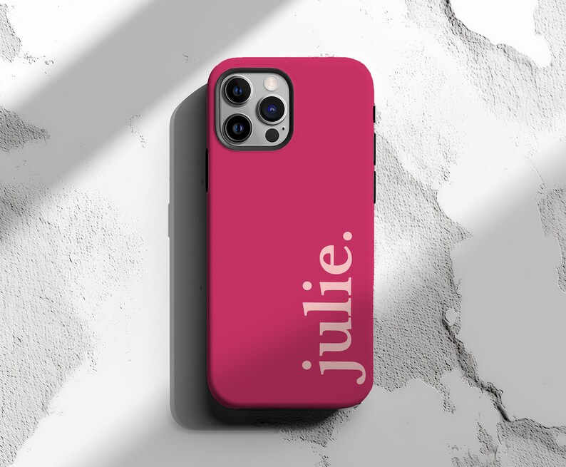 May include: A magenta phone case with the name "julie." printed vertically in a light color. The case has a camera cutout and a black border. The background is a textured white surface.