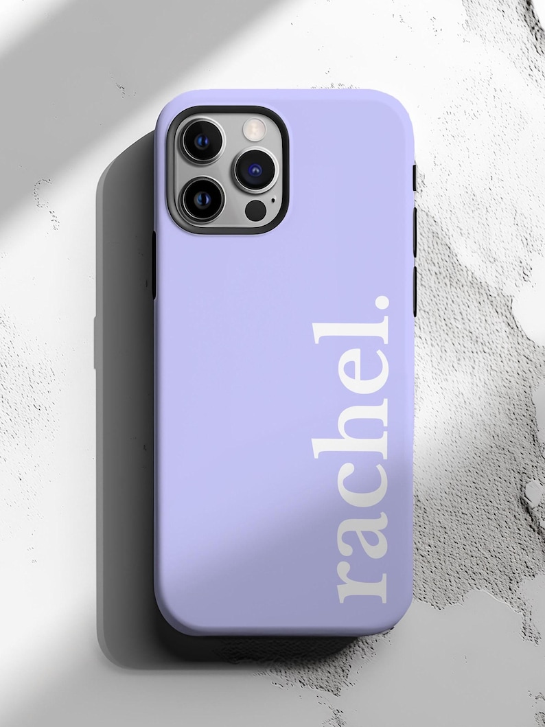 May include: A lavender phone case with the name "rachel." printed vertically in white. The case has a black camera module and buttons on the side. The phone case is on a white and gray textured surface.