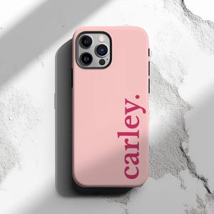 May include: A light pink phone case with the name "carley." printed vertically in a darker pink font. The case has a black camera housing and buttons. The background is a textured white surface.