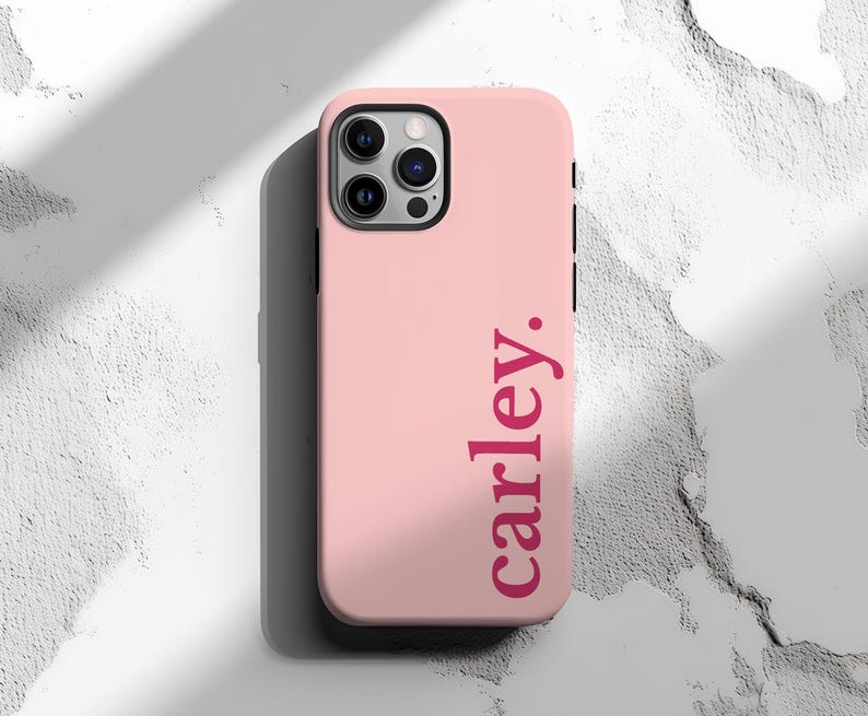 May include: A light pink phone case with the name "carley." printed vertically in a darker pink font. The case has a black camera housing and buttons. The background is a textured white surface.