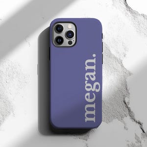 May include: A periwinkle phone case with the name "megan." printed vertically in white. The case has a camera cutout and is designed for a smartphone. The background is a textured white surface.