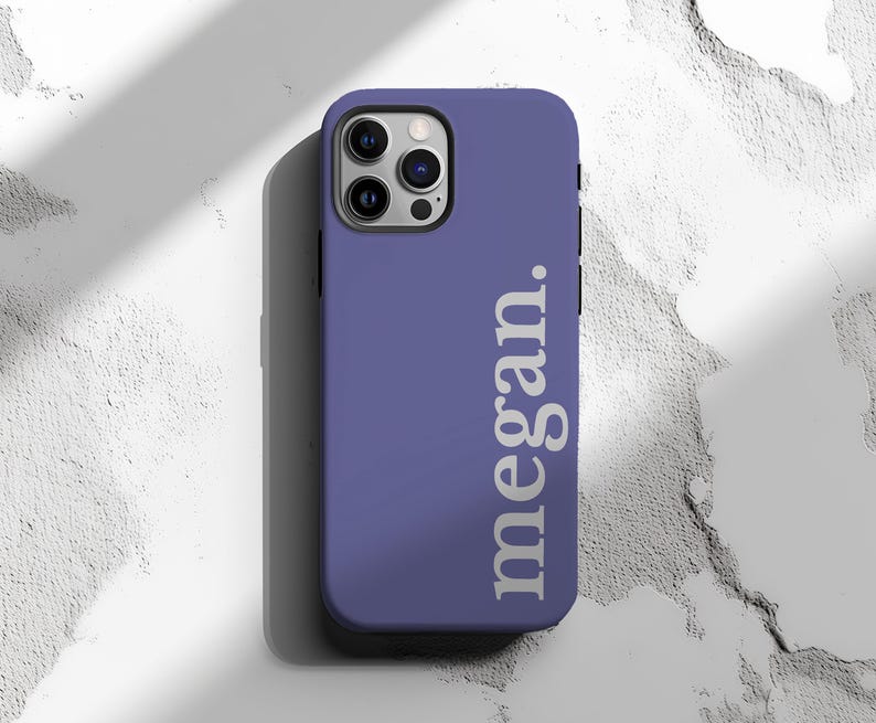 May include: A periwinkle phone case with the name "megan." printed vertically in white. The case has a camera cutout and is designed for a smartphone. The background is a textured white surface.