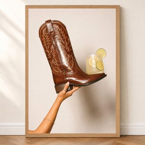 Peut inclure: Une botte de cow-boy en cuir marron avec des motifs de surpiqûres blanches complexes sur la tige, surmontée d'un verre de boisson citronnée jaune pâle avec deux tranches de citron vert sur le bout, le tout dans un cadre en bois clair.