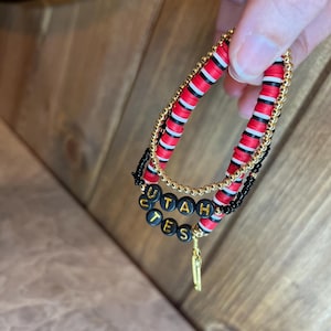 May include: Three layered bracelets with black and red beads. The top layer is gold beads. The bottom layer has black beads with the word "UTAH" and "UTES" spelled out in gold letters.