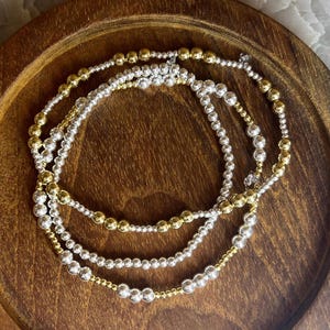 May include: Three layered bracelets with silver and gold beads. The bracelets are on a wooden surface.