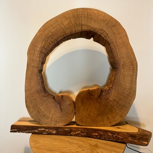 May include: A wooden lamp with a unique design featuring a large, circular, open-ended piece of wood with a smaller piece of wood on the bottom. The lamp is on a wooden table.