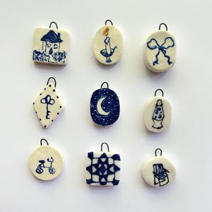 May include: Nine ceramic ornaments with blue and white designs. The ornaments feature a house, a candle, a bow, a key, a crescent moon, a lava lamp, a bicycle, a quilt pattern, and a chair.