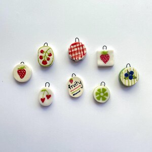 May include: A collection of nine small, round, and square ceramic charms with fruit designs. The charms feature strawberries, cherries, blueberries, and a lime. One charm has the word "fruity" written on it. Each charm has a small metal loop.