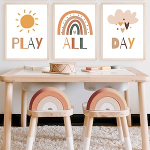 May include: Three framed prints with a sun, rainbow, and cloud with hearts. The prints have the words "PLAY ALL DAY" on them. The prints are hanging on a wall above a wooden table with two rainbow-shaped chairs.