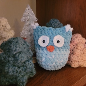 May include: A blue crocheted owl with white eyes and a red nose, surrounded by crocheted trees and a white flocked Christmas tree.