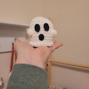 May include: A white crocheted ghost with black felt eyes and a mouth. The ghost is being held in a person's hand.