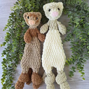 May include: Two handmade crochet animal toys. One is brown and tan, the other is cream and light green. Both have bear-like faces with pink noses and knotted feet. They are displayed against a backdrop of green foliage.