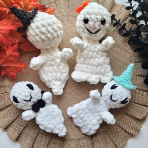 May include: Four white crocheted ghost ornaments with different facial expressions and accessories. One ghost wears a black witch hat, another has a red bow, another has a blue and green hat, and the last one has a black bow tie.