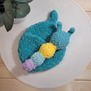 May include: A teal blue crocheted caterpillar toy with yellow, light blue, and purple accents. The toy is lying on a white crocheted placemat.