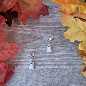 May include: Two small, white ghost charms with black dot eyes and silver-colored clasps. The charms are set against a backdrop of autumn leaves in shades of red, orange, and yellow, suggesting a Halloween or fall theme.