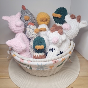 May include: A white wicker basket filled with crocheted farm animal plush toys. The basket contains a pink pig, chickens, ducks, and cows. The basket has a floral patterned liner and sits on a white woven placemat.