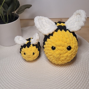 Peut inclure: Deux abeilles crochetées, une grande et une petite, avec des rayures noires et jaunes et des ailes blanches. Les abeilles sont assises sur un tapis circulaire blanc.