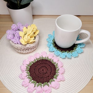 May include: Three crocheted coasters in pink, blue, and brown with a flower design. The coasters are on a white crocheted placemat.