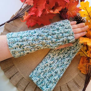 May include: A pair of teal, white, and green knitted fingerless gloves. The gloves are made with a cable knit pattern and are lying on a brown burlap surface.