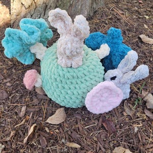 May include: A crocheted plush toy set featuring a green sphere with pink and blue crocheted flowers, a beige bunny sitting on top of the sphere, and a grey bunny with a pink ear.