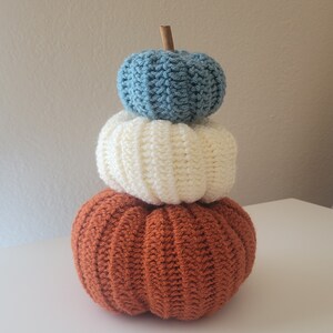 May include: A stack of three crocheted pumpkins in orange, white, and blue. The pumpkins are stacked on top of each other, with the smallest pumpkin on top.