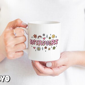 May include: White ceramic mug with a pink, yellow, and blue graphic that says "Introvert" surrounded by small flowers, stars, and hearts.