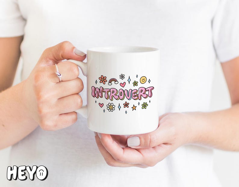 May include: White ceramic mug with a pink, yellow, and blue graphic that says "Introvert" surrounded by small flowers, stars, and hearts.