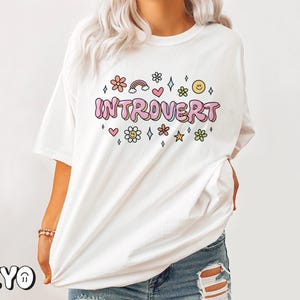 May include: White t-shirt with the text "Introvert" in pink letters surrounded by colorful flowers, hearts, stars, and a rainbow.