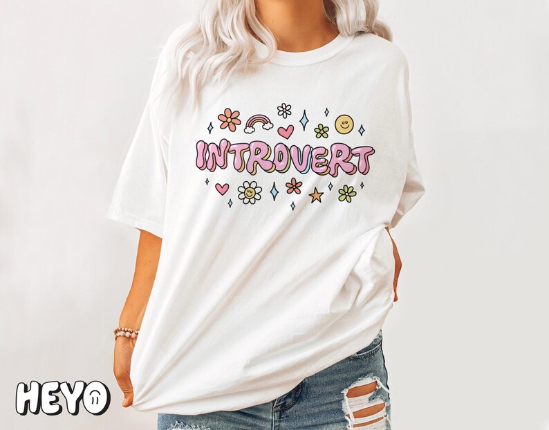 May include: White t-shirt with the text "Introvert" in pink letters surrounded by colorful flowers, hearts, stars, and a rainbow.