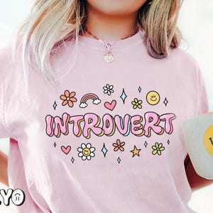 May include: Pink t-shirt with the text "Introvert" in pink bubble letters. The text is surrounded by small pink flowers, hearts, stars, and rainbows.