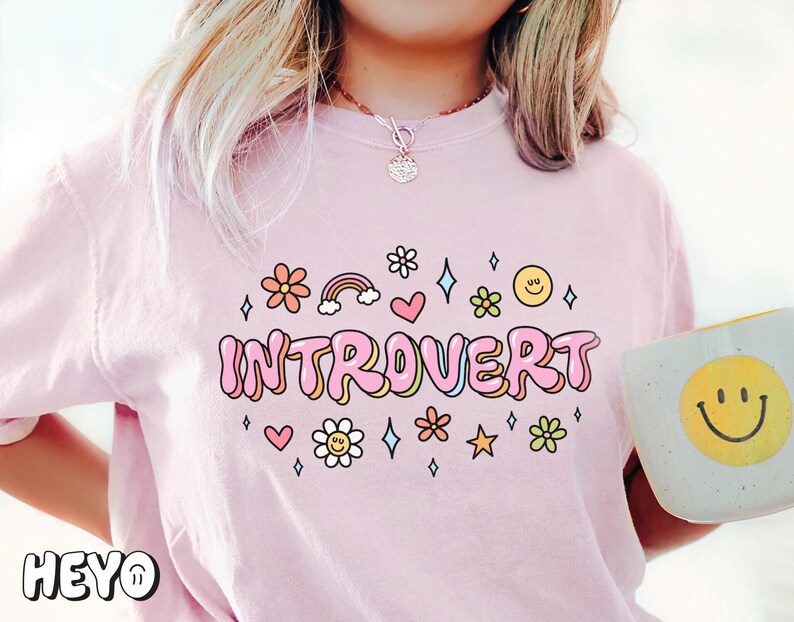 May include: Pink t-shirt with the text "Introvert" in pink bubble letters. The text is surrounded by small pink flowers, hearts, stars, and rainbows.