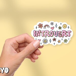 May include: A white sticker with pink lettering that says "Introvert" surrounded by colorful flowers, stars, and rainbows. The sticker is being held by a hand.