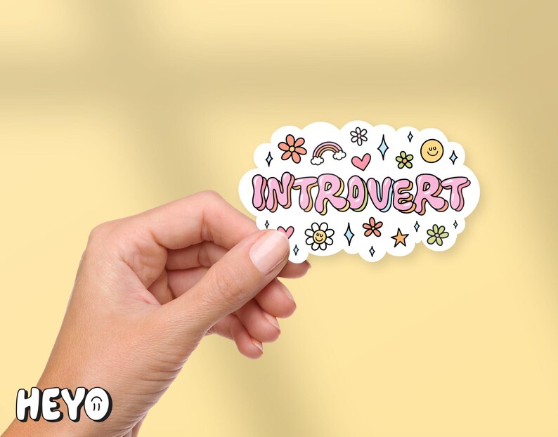 May include: A white sticker with pink lettering that says "Introvert" surrounded by colorful flowers, stars, and rainbows. The sticker is being held by a hand.