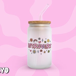 May include: White glass jar with a wooden lid and a straw. The jar has a pink and white design with the text "Introvert" and colorful flowers and stars.