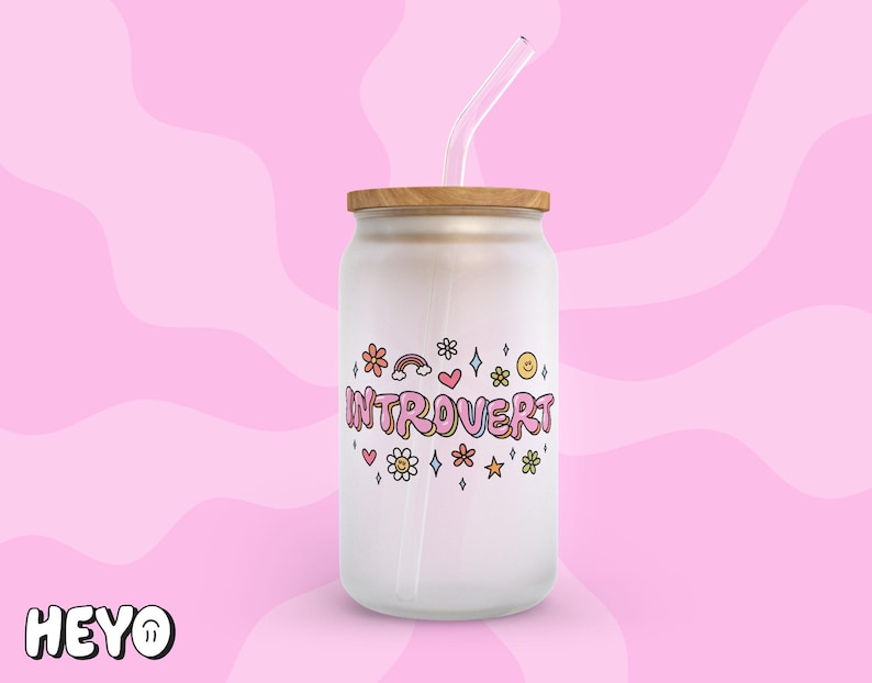 May include: White glass jar with a wooden lid and a straw. The jar has a pink and white design with the text "Introvert" and colorful flowers and stars.