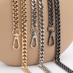 Puede incluir: Cuatro correas de cadena para bolso diferentes, oro, plata y dos tonos de negro, se muestran sobre una superficie de color marrón claro. Las cadenas son todas del mismo estilo, una cadena gruesa y robusta.