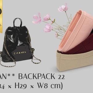 Puede incluir: Una mochila de cuero negro con cierre de cordón y herrajes dorados. La mochila mide 34 cm de largo, 29 cm de alto y 8 cm de ancho. La imagen también muestra un inserto de bolsa de fieltro beige, rosa y burdeos.