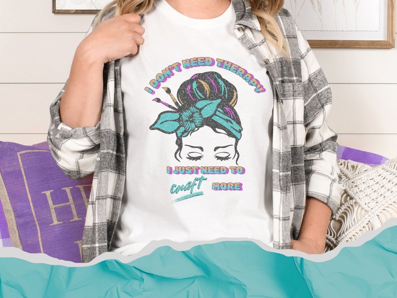 A white softstyle shirt that says I don't need therapy I just need to craft more. Graphic is of a woman with a messy hair bun and paint brushes sticking out of the headband. Colors in graphic and font are aqua, pink, orange and black.