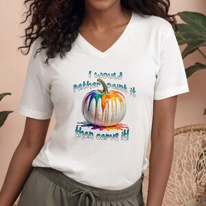 K&ouml;nnte beinhalten: Wei&szlig;es V-Ausschnitt-T-Shirt mit einem bunten, bemalten K&uuml;rbis-Design. Der K&uuml;rbis hat tropfende Regenbogenfarben. Der Text auf dem Shirt lautet: "I would rather paint it than carve it!"