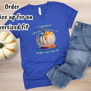 K&ouml;nnte beinhalten: Ein k&ouml;nigsblaues V-Ausschnitt-T-Shirt mit dem Text "I would rather paint it than carve it!" und einer Grafik einer wei&szlig;en K&uuml;rbis mit bunten Farben, die heruntertropfen. Hellblaue Jeans liegen neben dem Shirt.