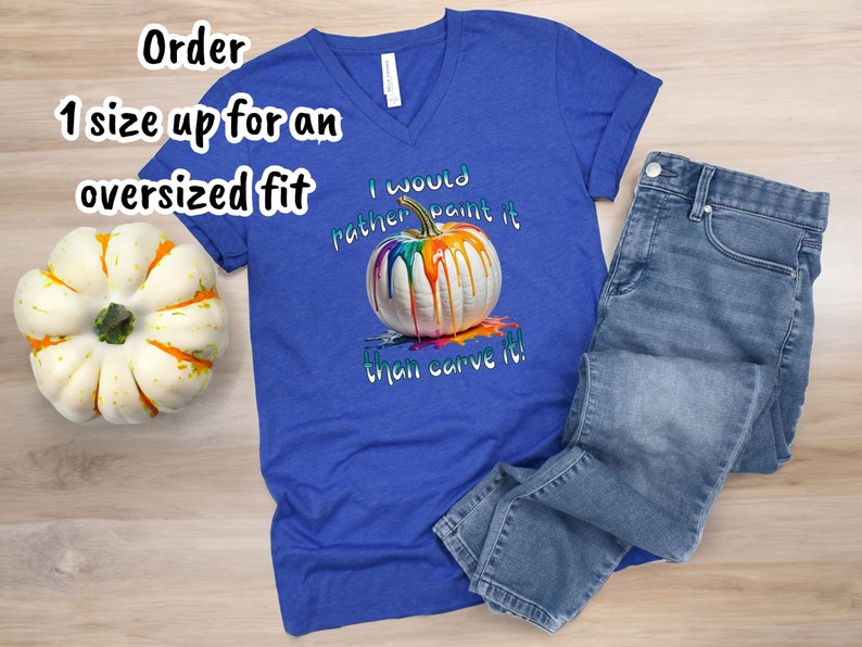 K&ouml;nnte beinhalten: Ein k&ouml;nigsblaues V-Ausschnitt-T-Shirt mit dem Text "I would rather paint it than carve it!" und einer Grafik einer wei&szlig;en K&uuml;rbis mit bunten Farben, die heruntertropfen. Hellblaue Jeans liegen neben dem Shirt.