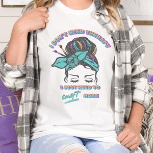 A white softstyle shirt that says I don't need therapy I just need to craft more. Graphic is of a woman with a messy hair bun and paint brushes sticking out of the headband. Colors in graphic and font are aqua, pink, orange and black.