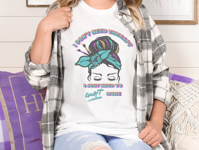 A white softstyle shirt that says I don't need therapy I just need to craft more. Graphic is of a woman with a messy hair bun and paint brushes sticking out of the headband. Colors in graphic and font are aqua, pink, orange and black.