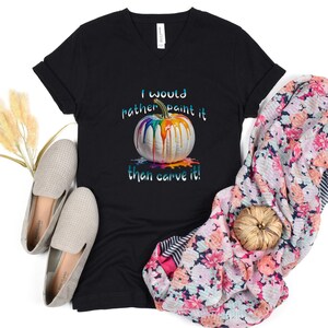 K&ouml;nnte beinhalten: Schwarzes V-Ausschnitt-T-Shirt mit dem Text "I would rather paint it than carve it!" und einer Grafik einer wei&szlig;en K&uuml;rbis mit bunten Farben, die heruntertropfen. Das Shirt wird mit einem floralen Schal und grauen Schuhen pr&auml;sentiert.