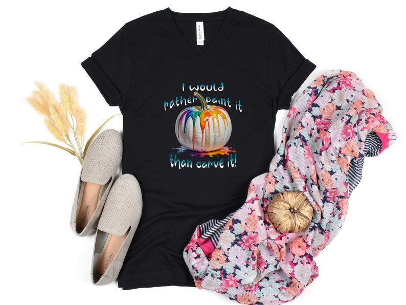 K&ouml;nnte beinhalten: Schwarzes V-Ausschnitt-T-Shirt mit dem Text "I would rather paint it than carve it!" und einer Grafik einer wei&szlig;en K&uuml;rbis mit bunten Farben, die heruntertropfen. Das Shirt wird mit einem floralen Schal und grauen Schuhen pr&auml;sentiert.